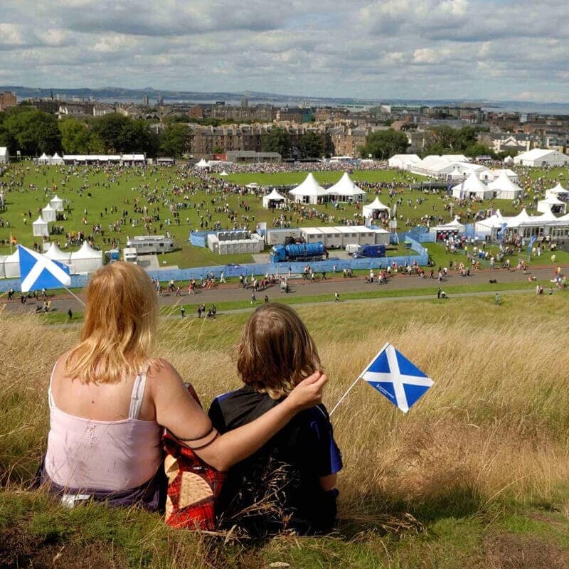 A photo of an woman and child holding small Scottish saltire flags, sitting on a hillside watching the crowds and tents of The Gathering 2009